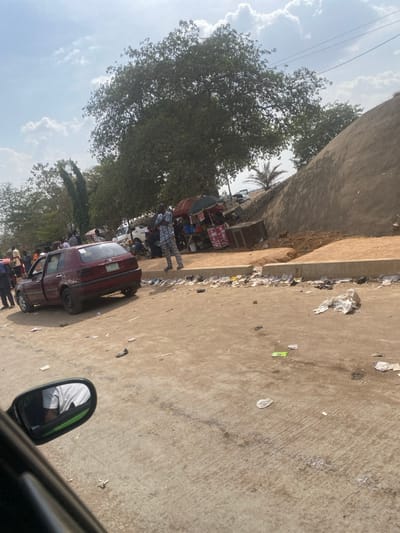 This image depicts an outdoor street scene in Iddah, Nigeria, under a bright, partly cloudy sky. The foreground shows a dusty, uneven road surface, possibly concrete or hardened earth, extensively littered with plastic bags, paper, and general refuse.
On the left side of the road, a dark red two-door hatchback vehicle, consistent with a Volkswagen Golf Mk3, is stationary, angled slightly away from the viewer. Its passenger door is open. A group of approximately 5-7 individuals is gathered near this vehicle.
On the right side of the road, a makeshift market stall is set up at the base of a steep, earthen mound. The stall consists of a counter or table beneath a red and white striped umbrella canopy, displaying various packaged goods. A male individual, dressed in a patterned blue and white outfit, stands with his arms crossed on a low concrete barrier adjacent to this market stall. Further into the background, a white pickup truck is partially visible behind the stall.
Beyond the road and structures, large trees with dense green foliage dominate the left and center-background. The side mirror of a vehicle, from which the photograph was taken, is visible in the lower left corner, reflecting the arm and torso of a person wearing a white shirt. The overall environment suggests a roadside area with commercial activity and a noticeable accumulation of debris.