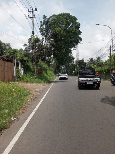The image depicts a rural road scene in Khajuripada, India, under a partly cloudy sky. The asphalt road, marked with a solid white line on its left edge, recedes into the distance. On the left side, a grassy and dirt shoulder leads to dense green vegetation, including overgrown bushes and several trees, one notably large with a full canopy. A wooden utility pole with multiple cross arms and insulators stands on the left, carrying numerous power lines. Adjacent to the road, behind the vegetation, is a dilapidated structure partially obscured by a brown corrugated metal fence or gate. A small red sign is attached to a slender tree trunk on the left.
In the mid-ground, a white sedan travels away from the viewer, positioned toward the center of the road. In the foreground, a black pickup truck with a utility bed also travels away from the viewer, positioned on the right side of the road. Its tailgate features colorful stickers and the license plate "B 9054 HI" is visible. A red object is present in the truck's bed. On the far right, partially obscured by the pickup truck, a person on a dark scooter or motorcycle, wearing a helmet, is also traveling in the same direction. The right side of the road is lined with more dense greenery, including visible palm trees further back, and features another utility pole extending a street light arm over the road, carrying a multitude of tangled black wires. A tall, metal lattice communication tower is visible in the distance behind the trees on the right.
