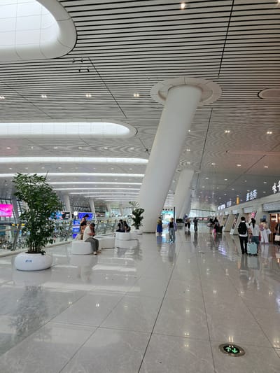 This image captures the interior of a modern transportation hub, likely a train station or airport, in Shangcheng District, China. The scene is characterized by a vast, brightly lit space with a high, horizontally-slatted ceiling featuring recessed lighting. Large, white, cylindrical pillars support the ceiling, one of which is prominently angled in the foreground.
The floor is highly polished and reflective, appearing to be large, light-colored tiles. In the mid-ground, several individuals are visible. Some are seated on minimalist white benches, appearing to rest or wait. Others are standing and walking, with a few individuals pulling luggage, indicating travel. A large potted plant with green foliage is situated in the left foreground, adding a touch of nature to the otherwise sterile environment.
In the background, digital displays and signage are visible, suggesting areas for information or retail. The overall impression is one of a clean, spacious, and efficiently designed public space. The architecture is contemporary and the lighting is designed to create an airy and open atmosphere. The presence of people with luggage and directional signs confirms the function of the location as a place of transit.