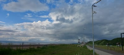 The image depicts a coastal scene in Aleksandrovsk-Sakhalinskiy, Russia, likely during overcast weather, with a view of the Sea of Okhotsk on the left. A dark, sandy beach runs along the shoreline, bordered by a grassy area in the foreground. A paved pathway runs parallel to the beach on the right side of the image, with several lampposts, some equipped with cameras, lining the path. In the middle ground, there is a small playground with a slide and a lifeguard-like tower. Beyond the playground, a line of green hills rises, dotted with a few vehicles parked in a grassy field. The sky is filled with a dramatic mix of dark, heavy clouds and patches of bright blue sky. No people are visible in the scene.