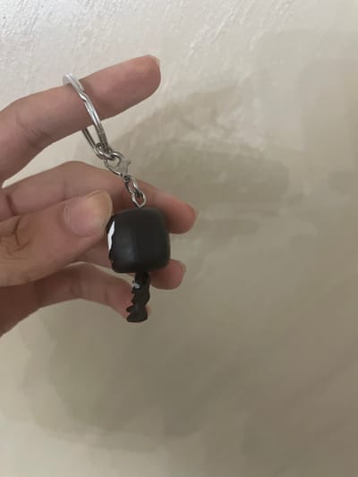The scene is located in the city of Ihtiman, Bulgaria. A light-skinned human hand holds a keychain vertically against a plain, light-colored wall. The keychain features a silver-toned split key ring positioned on an index finger, a small metal chain, a lobster clasp, and a miniature character charm. The charm is predominantly black, with a large, squared head featuring a distinctive white marking on its left side, and a smaller, black body with defined legs below. The hand firmly grips the keychain components. The background wall displays minor textural variations and diffuse lighting.