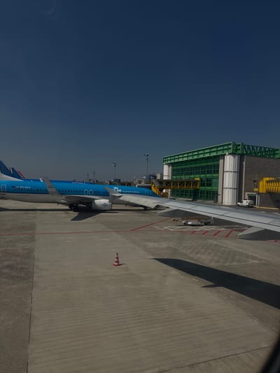 The image depicts an airport scene in Peschiera Borromeo, Italy, featuring KLM Royal Dutch Airlines aircraft parked at a terminal. The aircraft's blue livery is visible. The airport terminal building, with green-framed windows, runs parallel to the aircraft. An orange and white striped safety cone stands on the apron. The sky is clear and blue.