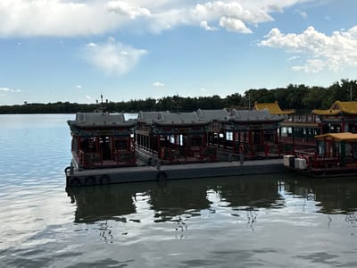 The image displays a tranquil scene on a body of water in Haidian District, China. A pale blue sky, accented with scattered white cumulus clouds, occupies the upper portion of the frame. Below, a distant, flat shoreline is densely covered with dark green trees, forming a continuous band across the midground. The water surface, a muted blue-grey, shows subtle ripples and reflects the sky and the structures along its edge.
In the foreground and midground, a dark grey floating dock, equipped with black cylindrical fenders along its side, extends horizontally. Moored alongside this dock are multiple traditional Chinese-style boats. Several boats feature distinctive grey, tiled, upturned roofs with intricate green, red, and gold decorative elements along their eaves and railings. The main bodies of these boats are predominantly red-brown with visible window openings and railings. Towards the right side of the dock, at least two boats are visible with prominent bright yellow, similarly styled, upturned roofs. Orange life rings are discernable on the railings of some boats. Reflections of the boats and the dock are clearly visible on the water's surface beneath them. No people are present on the boats or the dock.