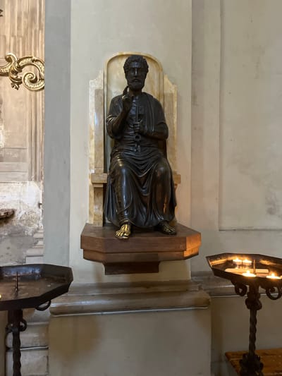 A bronze sculpture of Saint Peter with keys rests on a marble base and a wooden stand in a church setting in Bologna, Italy. The sculpture is positioned against a neutral wall with decorative trim. Nearby are ornate metal candle holders. The background includes architectural details of the church, suggesting an indoor, religious environment.