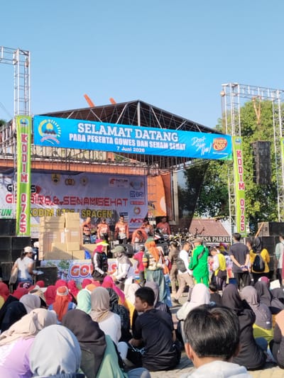 This image depicts an outdoor community event in Sragen, Indonesia, on a clear, sunny day, dated June 7, 2026. A large, temporary metal truss stage is central, adorned with multiple banners. The prominent horizontal blue banner across the top reads "SELAMAT DATANG PARA PESERTA GOWES DAN SENAM SEHAT" (Welcome to the participants of Cycling and Healthy Exercise), featuring the crest of "KABUPATEN SRAGEN" (Sragen Regency) and logos for "Hydro Coco" and "Don't Stop Now!". Vertical green banners on either side of the stage display "KODIM 0725" (Military District Command 0725), with the text "KODIM 0725 / SRAGEN" also visible on a lower banner. The stage backdrop includes sponsor logos such as "FASTLINK INTERNET PROVIDER," "BRI," "ELITE Cell," "NSS," and "Polytron."
On the stage, approximately six to eight individuals, predominantly men, are visible; some wear matching orange and black shirts. Large stacks of cardboard boxes, some bearing the "Polytron" logo, are positioned on the left side of the stage, likely prizes or supplies. Sound speakers are mounted on the truss structure on either side.
In the mid-ground and foreground, a substantial crowd is gathered, with many individuals seated directly on a paved ground surface. The audience consists of diverse age groups, with a notable presence of women wearing hijabs and men in casual attire. Several people stand in the mid-ground, observing the stage, including a man in a bright green tracksuit walking towards the right. In the immediate foreground, the back of a seated man's head, wearing a black t-shirt and grey hoodie, is prominent. The background reveals scattered trees and glimpses of distant residential or commercial buildings, including a sign for "ESA PARFUM," under a light blue sky, indicating an open public space within the city of Sragen.