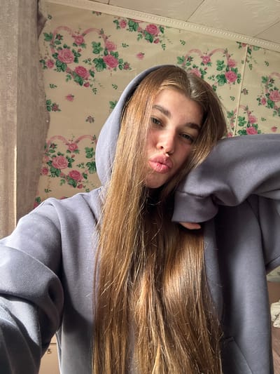 This media file shows a close-up, vertically oriented selfie of a young woman with long, light brown hair. She is wearing a light purple hoodie, with the hood pulled up around her head. Her face is turned towards the camera, and she is making a kissing or puckering expression with her lips. Her eyes are open and looking at the camera, with blue irises. Her eyebrows are dark and well-defined.

The background consists of wallpaper with a floral pattern of pink roses and green leaves on a cream-colored base. There is also a textured white trim running along the top edge of the visible wallpaper. The lighting suggests it is daytime, possibly with sunlight casting shadows, creating a warm glow on her hair and face.

The context provided indicates the location is Мещёры, Russia. There is no visible text in the image. The overall mood conveyed by the selfie is playful and casual.