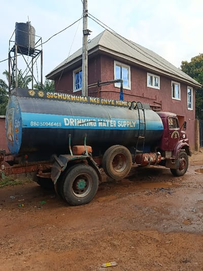 A maroon water truck sits on a muddy road. "Drinking Water Supply" is painted on the blue tank, along with other words and numbers. A water tower stands to the left of a two-story brown house with a tan roof and white-framed windows. Utility poles with wires are also visible. The scene appears to be in a rural area.