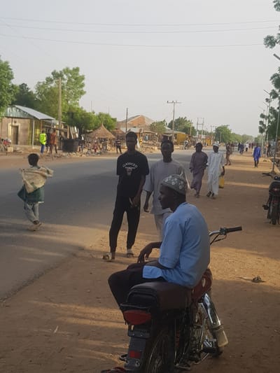 The image shows a group of people gathered around a motorcycle on a street in Dabawa, Nigeria. A man in a light blue shirt is bent over the motorcycle, appearing to be working on it. Two other men stand nearby, observing. One man is wearing a black t-shirt and the other is wearing a white kufi. Other individuals are seen walking along the street in the background, some dressed in traditional clothing. The environment includes buildings, trees, and power lines lining the street. A motorbike is parked in the distance.