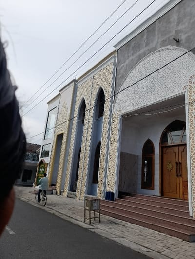 The image shows a mosque located in the city of Kecamatan Tanggulangin, Indonesia. The mosque is a large, modern structure with a gray facade and intricate white designs on the exterior. It has three entrances and a set of stairs leading up to the main entrance. The building appears to be freshly painted.  The mosque is located on a street with a paved sidewalk in front. A man is riding a bicycle on the street, and a small trash can is visible on the sidewalk. The sky is clear and blue with a few light clouds.  The coordinates of the location are -7.504 latitude and 112.715 longitude. The mosque is likely a place of worship for Muslims in the area.