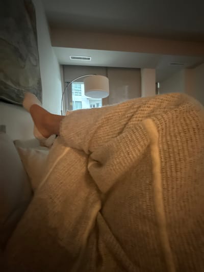 The image depicts an indoor scene, likely a residential unit or hotel room, within an urban environment in the United States; a precise location within Washington cannot be determined. The primary subject is a partially visible person reclining, with their left leg elevated, bent at the knee, and resting on a white pillow. The foot is covered by a white sock. The lower leg and torso are covered by a light beige, textured, knitted blanket or garment. In the background, an abstract artwork with gray and white patterns hangs on the wall. A modern arc floor lamp with a white drum shade stands to the right of the artwork, providing ambient illumination. A window reveals a portion of an exterior multi-story building. A ceiling vent is visible in the upper background. The overall lighting is dim, suggesting evening or low ambient light.