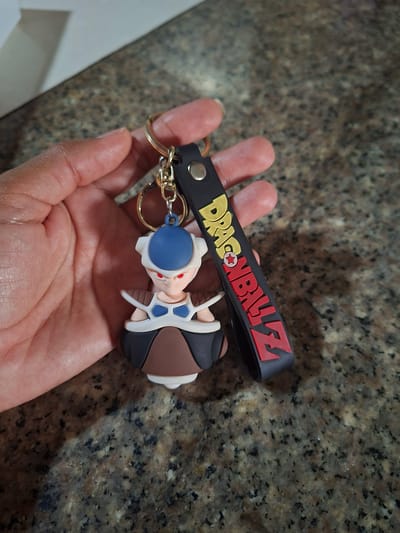 A person's hand is holding a keychain with a Dragon Ball Z charm. The charm is of a character with a blue helmet, red eyes, and white and brown armor. The keychain also has a black strap with the words "DRAGON BALL Z" written in yellow and red. The background is a speckled granite countertop. There is no information available to indicate the location of El Valle del Espíritu Santo, Venezuela.