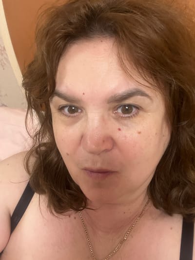 This is a close-up selfie of a middle-aged woman with brown hair and brown eyes. She is wearing a black bra and a gold chain necklace. The image is taken indoors, in what appears to be a bedroom. The background shows a wall with a subtle floral pattern and a corner of a brown headboard or wall panel. There are no discernible landmarks or indicators to suggest the location is Tula, Russia. The woman has a few moles on her face and neck.