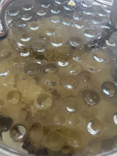 The image shows a close-up view of condensation droplets forming on the underside of a clear pot lid, which is covering food. The food appears to be a pale yellow, possibly mashed potatoes or a similar dish, with some darker, indistinct pieces mixed in. The condensation droplets are numerous and vary in size, some appearing as perfect spheres, while others are elongated or irregular. Light reflects off the curved surfaces of these droplets, creating bright highlights. The lid itself is likely glass or a clear plastic, and a metal handle or edge of the pot is visible on the left side of the frame. The scene suggests a cooking or steaming process. The image does not provide any information about a specific location in Richmond Hill, Canada, or any people, actions, or interactions.