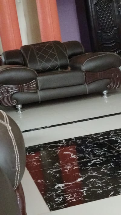 The image captures an indoor living space, likely a living room, featuring a dark brown two-seater sofa as its central subject. The sofa, possibly upholstered in leather or faux leather, is seen from a slightly elevated angle, revealing its detailed construction. Its back cushions feature a distinctive diamond-patterned stitching, and the armrests are adorned with ornate, carved wooden designs in a dark reddish-brown hue. The sofa stands on small, shiny, metallic feet.
In the bottom left foreground, a partial view of another similar dark brown sofa or armchair is visible, showcasing part of its armrest with prominent white stitching.
The flooring consists of light-colored, possibly white or off-white, tiles in the midground, with a striking section of black marbled tiles featuring white veining in the foreground. This dark, patterned area suggests either a decorative rug or a distinct tile layout.
The background reveals vertical elements that appear to be drapes or curtains in varying colors: orange/peach on the left, a lighter neutral shade, and a section of purple towards the right. To the far right, a dark, intricately carved wooden door or large furniture piece with a patterned surface is partially visible.
No people or specific activities are depicted, giving the scene a static and observational quality. The lighting is ambient, characteristic of an indoor setting, and no visible text is present. The overall aesthetic suggests a traditionally or semi-traditionally decorated interior, consistent with a home or formal reception area. The provided location context is Abakaliki, Nigeria.