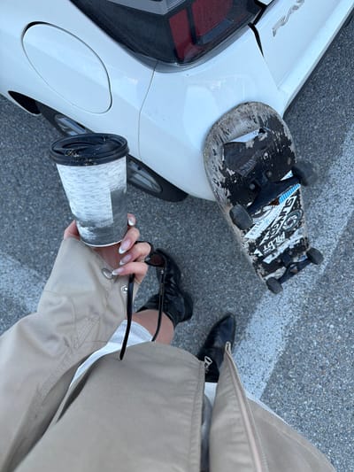 A high-angle medium shot shows a person standing beside a white vehicle, holding a coffee cup and sunglasses. A worn skateboard rests on the vehicle's surface. The person wears black boots and a khaki-colored jacket. The location is identified as Toseong-myeon, South Korea.