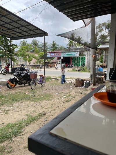 The image depicts a street scene in Cotseutui, Indonesia, under an overcast sky. In the foreground, a portion of a table with a tiled surface is visible, on which rests an orange saucer with a glass of dark liquid, likely coffee. To the left of the table, a dirt and grass area is shown, with scattered debris.
Dominating the mid-ground is a dark motorcycle parked on the side of the road. Next to it is a small purple bicycle with a woven basket on the handlebars. A rooster is positioned near the front wheel of the motorcycle.
Further back, a young girl in a white t-shirt and blue jeans stands with her hands behind her back, looking towards the right. Behind her, a street runs horizontally, with several buildings lining its sides. One building displays a sign reading "DR. Kupi" and another "UD. HARAPAN JAYA." Green metal gates and parked motorcycles are visible in front of these establishments. Palm trees and other vegetation are scattered throughout the scene, suggesting a tropical or subtropical climate. The overall atmosphere is casual and appears to be a typical street view in a populated area of Indonesia.