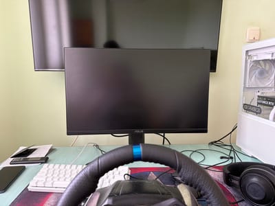 The image captures an indoor gaming and computer setup, likely in a room with light green walls. The scene is dominated by a desk featuring various electronic devices and peripherals.

In the foreground, a black gaming steering wheel with a distinctive blue stripe at its top center is prominently placed, suggesting a focus on racing simulation games. Behind the steering wheel, a white mechanical keyboard with visible keys like "Caps Lock" and "Shift" is situated. A black smartphone, some papers, a black card, and a small black electronic device are arranged on the left side of the desk.

Centered on the desk is a black ASUS monitor, currently displaying a dark, unlit screen. Above and slightly behind it, a larger, wall-mounted dark monitor is partially visible. To the right, a white desktop PC tower stands on the desk, featuring a transparent side panel that showcases internal components, including a white fan and a graphics card with "GEFORCE RTX" text. The PC case itself has "GRIGS" printed on it. A pair of black Razer headphones rests nearby. Numerous black cables are visible, running across the desk and behind the monitors.

On the light green wall to the right, a beige wall-mounted switch or control panel is visible. The overall setting is a personal computing station, likely used for gaming, depicted during what appears to be daytime, given the general lighting. No specific activity is occurring, and no people are present, thus no emotions are conveyed. The provided location context is Saedinenie, Bulgaria.