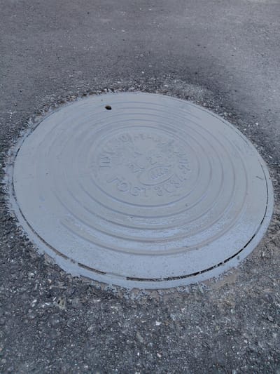 The image displays a close-up, slightly angled view of a light gray, round manhole cover set into dark asphalt pavement. The manhole cover appears recently painted or wet, with some lighter residue visible on its surface and edges where it meets the asphalt.
**Subjects:** The primary subject is a metal manhole cover. The surrounding material is asphalt pavement.
**Objects:** The main object is the manhole cover itself. The textured surface of the asphalt is also prominent.
**Scene, Setting, and Activity:** The scene is an outdoor ground-level utility access point, likely on a street or pathway, given the asphalt. There is no discernible activity taking place; it is a static shot.
**Notable details:**
- **Time of day/Weather:** The lighting suggests natural daylight, possibly overcast or diffused, as there are no strong shadows. The wet appearance of the manhole cover could indicate recent rain or a fresh coat of paint.
- **Location cues:** The text on the manhole cover provides a strong location cue to Russia due to the use of Cyrillic script and the GOST standard.
**Visible text:**
Embossed on the manhole cover, the following text is visible:
- "ГОСТ 3634-2019" (GOST refers to a set of technical standards in Russia and other post-Soviet states, with 3634-2019 likely indicating the specific standard and revision year for manhole covers).
- "ТК" (an abbreviation, possibly for a company or technical specification).
- An emblem or logo, which appears to be a stylized "Им" or "HM" within an oval shape.
- Faint numbers "10-1" are partially visible near the top left.