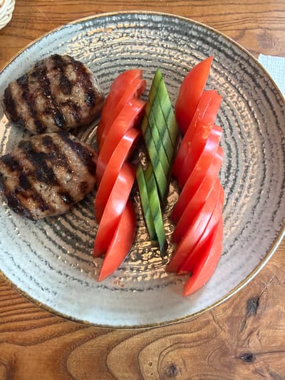 A top-down, close-up shot reveals a plate of food on a wooden surface, likely a table. The plate is round, with a gray and white patterned interior and a gold-colored rim. On the plate are two small, round, grilled patties exhibiting grill marks. Above the patties, sliced tomatoes are arranged in a row, topped with cut, skin-on cucumbers, also in a row. The wooden table displays natural grain patterns. The image suggests the food was served at a restaurant, possibly in Rudartsi, Bulgaria, though no specific elements in the image confirm the location directly.