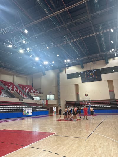 This is a wide-angle, slightly tilted shot of a basketball court inside a large indoor arena. The scene is lit by bright overhead lights, indicating it could be daytime or nighttime.

The primary subjects are a group of people, likely a basketball team, gathered in a huddle on the wooden court. Most of them are wearing athletic attire, with some in jerseys and shorts. They appear to be engaged in a pre-game or halftime discussion. There's a basketball hoop visible in the background, along with a digital scoreboard showing "HOME 89" and "GUEST 25", suggesting a game is either in progress or has recently concluded.

The arena features tiered seating sections with red and white chairs. banners and signs are visible on the walls and seats. One banner reads "FIBA YOUTH DEVELOPMENT FUND PROJECT". Another sign displays "ÇIKIŞ 5" and "ÇIKIŞ 7". The overall atmosphere seems focused and possibly tense, given the context of a basketball game. The polished wooden floor of the court reflects the bright lights.