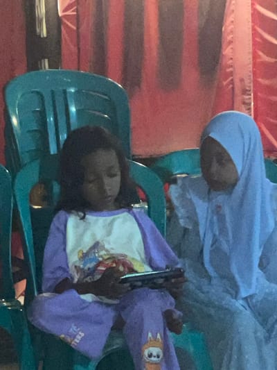 Two young girls are seated on teal plastic chairs, facing forward. The girl on the left, with dark hair, is wearing a purple and white long-sleeved shirt with graphics and purple pants. She is looking down at a smartphone she is holding in her hands. The girl on the right is wearing a light blue hijab and a light blue garment. They are seated in an indoor setting with a red backdrop. The image appears to have been taken in Wedarijaksa, Indonesia.