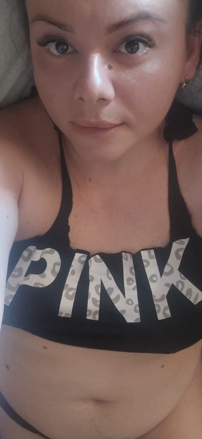 A close-up, vertically oriented, selfie-style photograph features a woman lying on her side, facing the camera. Her head is tilted and angled towards the viewer, with her right eye prominently in focus. She has dark hair and is wearing makeup, including mascara.
She is wearing a black sports bra or tank top with the word "PINK" printed across it in a white leopard-print pattern. The lower portion of the image reveals her midsection, including her navel. Her skin tone is fair, and there are a few small moles visible on her torso. The background is blurred and appears to be a soft, gray surface, possibly a bed or couch. The lighting suggests an indoor setting, possibly during the daytime given the natural-looking illumination on her face. There are no clear indications of time of day or weather. The overall mood conveyed is casual and relaxed. No specific activity is clearly depicted, other than posing for the photograph. The image is cropped in a way that focuses tightly on the woman and her upper body.