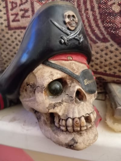 A close-up, eye-level, medium shot captures a resin sculpture of a skull wearing a black pirate hat adorned with a skull and crossbones emblem. The skull has a single green glass eye, a black leather eye patch inscribed with "Bermuda" in gold lettering, and visible teeth. The background consists of a patterned textile. The scene is set on a white shelf. The exact location in Aktau, Kazakhstan, is not determinable from the image.