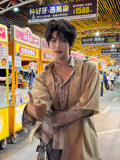 The image features a young person posing outdoors, likely at a night market in Guta, China. The background is densely adorned with strings of warm, yellow lights, creating a festive and illuminated atmosphere typical of evening street markets. Several stalls with colorful signage are visible, some displaying prices and product names in Chinese characters. One prominent sign reads "种好牙·选美康:1588" which translates to "Good teeth, choose Meikang: 1588," suggesting a dental service or product promotion. Another sign advertises "鸡蛋仔" (egg waffles) with prices listed.
The person in the foreground is wearing a layered outfit consisting of a plaid shirt and an open, light-colored jacket. They are looking towards the camera with a slight smile, and their right hand is raised in a gesture that could be interpreted as a thumbs-up or a peace sign. The overall mood appears relaxed and informal, capturing a moment of leisure amidst the bustling market. The time of day is clearly evening or night due to the artificial lighting dominating the scene. The ground visible in the lower part of the image appears to be a paved street, and a few wheeled carts are present, typical of market vendors.