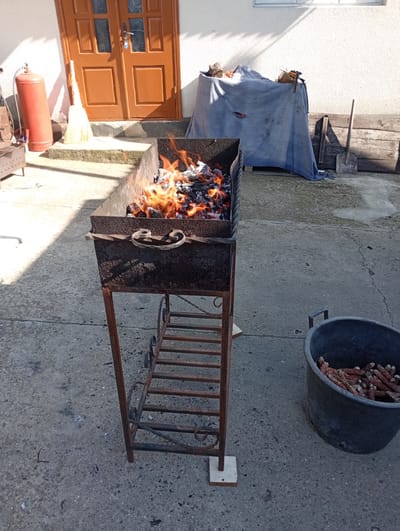 In Buțeni, Moldova, a courtyard scene unfolds under bright sunlight. The foreground features a rustic, rusty metal barbecue grill filled with burning embers and flames, suggesting recent or ongoing activity. To the right of the grill, a large, dark plastic tub is filled with dried corn cobs. In the background, a set of double wooden doors, painted brown, stand open, revealing a glimpse of an interior. To the left of the doors, a tall, red gas cylinder is positioned near a broom. Adjacent to the doors, a table is draped with a large, blue cloth, under which various objects are obscured. Further to the right, two wooden planks are stacked, and a shovel leans against them. The ground is paved with concrete. The overall impression is one of an outdoor, utilitarian space, likely part of a rural or suburban property.