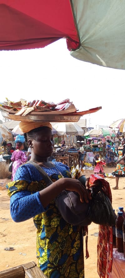 This image captures a vibrant, bustling outdoor market scene in Ketu Adieowe, Nigeria, under bright daylight.

The central figure is a woman, likely a vendor, positioned in the foreground and slightly off-center. She is dressed in a distinctive blue outfit featuring a patterned fabric with yellow, green, and brown motifs, topped with a blue lace-like material on the upper body. A golden chain with a cross pendant is visible around her neck. On her head, she skillfully balances a large, silver-toned metal tray, laden with numerous small, brightly colored packaged items, possibly snacks or sachets of food products, supported by a yellow headwrap. She also carries a dark shoulder bag, and from her arm or the bag hangs a black plastic bag containing multiple red-packaged goods. Her expression is focused, hinting at the demanding nature of her work.

The background showcases the lively market environment. The ground is sandy or dusty. Numerous market stalls are visible, characterized by their diverse coverings, including large white and colorful umbrellas and makeshift canopies, providing shade from the sun. Many other people, a mix of vendors and shoppers, are present throughout the market, engaging in daily commerce. Specific items on display at stalls include rows of bottled liquids (possibly palm oil or beverages) on the right side of the image, and a wooden stand in the mid-ground holding a collection of dark, round items, potentially food products. A notable detail is a white sign with the letters "POS" (Point of Sale) visible on a stall in the distance, indicating the availability of electronic transaction services. The overall atmosphere is one of dynamic activity and commerce typical of a busy African marketplace.