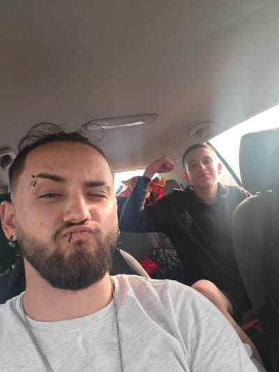 Two people are in a car. A man with a beard, lip piercing, and eyebrow piercing is in the foreground, making a kissing face and winking. Behind him is a younger person flexing their right arm. The interior of the car is visible, including the ceiling, seats, and other interior elements. The location is Bogdanovo, Bulgaria.