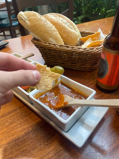 A close-up shot captures a hand dipping a tortilla chip into a white rectangular dish filled with a vibrant orange salsa, set against a wooden table. The chip, already partially submerged, is angled to scoop up the salsa. Adjacent to this dish, another smaller white dish holds a few green olives. In the background, a woven basket overflows with two long, lightly browned bread rolls sprinkled with sesame seeds, and a crumpled white napkin. To the right, the neck and label of a dark glass beer bottle are visible. The environment suggests an indoor or semi-outdoor dining setting with diffused natural light illuminating the scene.