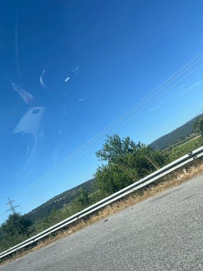 The image shows a view from a moving vehicle, likely on a road with a guardrail. The sky is a clear, bright blue with some wispy white clouds and reflections from the vehicle's interior, including a dash cam. In the distance, there are rolling green hills dotted with buildings, suggesting a rural or semi-rural area. Power lines run diagonally across the upper portion of the image. A metal guardrail runs along the side of the road, separating it from the vegetation and landscape beyond. The road surface appears to be asphalt. The overall impression is a snapshot of a journey through a landscape under a clear sky. The location is identified as Νέα Μεσολακκιά, Greece, implying a specific geographical context for this roadside scene.