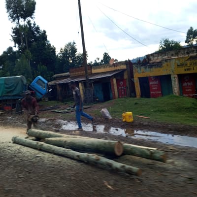 The scene depicts a street in Borabu Chitago ward, Kenya, after rainfall, with puddles on the unpaved ground. In the foreground, three logs lie amidst sawdust, indicating recent cutting activity; a man operating a chainsaw is visible on the left. Another man is walking past the logs, away from the chainsaw operator.
Behind them, a truck with a blue cab sits askew. The backdrop features a row of dilapidated buildings with advertisements for "Airtel Money Hapa" and "Wines &". The buildings are painted in yellow and red, but the paint is peeling. Beyond the buildings, trees are visible. The sky is overcast, suggesting recent or ongoing rain.