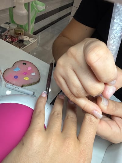 The image depicts a nail care session occurring at a salon in Naples, United States. Two individuals are present: a client, exhibiting lighter skin tones, and a nail technician, whose arm and hand show a slightly darker skin tone, wearing a black top. The technician's hands are actively engaged with the client's left hand. Specifically, the technician's right hand is manipulating the client's left ring finger, which has a clear, elongated nail. The client's left index finger features a clear nail decorated with a white, pink, and yellow floral pattern.
A pink and white UV/LED nail lamp is positioned beneath the client's left palm. On the white table surface to the left, a heart-shaped pink palette contains distinct drops of green, light blue, pink, purple, yellow, white, red, and black nail polishes. Adjacent to the palette are a dark brown nail art brush and two white cylindrical tools. In the background, a white utility cart holds various small jars, containers, and a white spray bottle. A green plastic bag hangs from the cart. The floor behind the cart displays dark horizontal stripes.