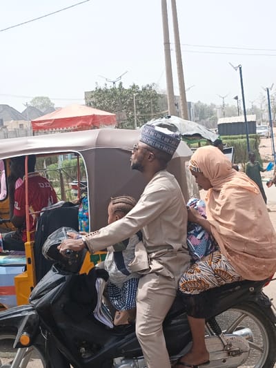 In Hotoro South, Nigeria, a family is observed on a black motorcycle in daylight. A man, wearing a light-colored long-sleeved tunic, matching trousers, a patterned blue and white cap, and sunglasses, operates the motorcycle, holding the handlebars. A young child with braided hair is positioned in front of the man, between him and the handlebars. Behind the man, a woman is seated as a passenger, wearing a light brown hijab, a patterned multi-colored skirt, and black footwear. She holds a patterned item or bag.
To the immediate left and slightly behind the motorcycle, a yellow and brown auto-rickshaw (Keke Napep) is partially visible, containing luggage and at least one male passenger in a red shirt. The background features include utility poles with multiple wires, streetlights equipped with solar panels, a red and white canopy, various buildings with residential-style roofs, scattered trees, and other individuals. The ground surface appears to be a mix of paved and unpaved areas. The sky is bright and overcast, suggesting an outdoor urban or semi-urban environment.
