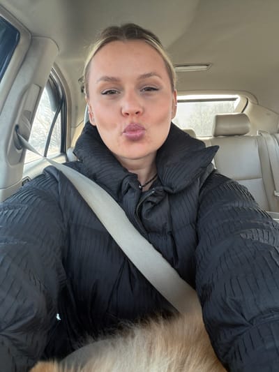 The image displays a female individual situated inside a vehicle, captured in a selfie perspective. She is positioned in what appears to be the driver's seat, secured by a light-colored (beige/tan) seatbelt diagonally across her chest. Her light-colored hair is pulled back, revealing a clear complexion with light makeup, including eyeliner. She wears small stud earrings and a thin black beaded necklace. Her lips are pursed forward in a distinct "kissing" expression, and she is looking directly at the camera. Her attire consists of a black, high-collared puffer jacket with a prominent textured, ridged pattern. In the lower foreground, partially visible below the seatbelt and jacket, is a mass of light-colored, fluffy fur, consistent with an animal, likely a dog. The vehicle's interior features light-colored (beige/tan) upholstery on the rear seats and headliner. Through the left-side window, bare tree branches are visible against a light, hazy sky, indicating an outdoor setting, possibly during colder months. The rear window shows a similar light sky. The specific geographic location in Дулесово, Russia, is not discernible from any visual cues within the image.
