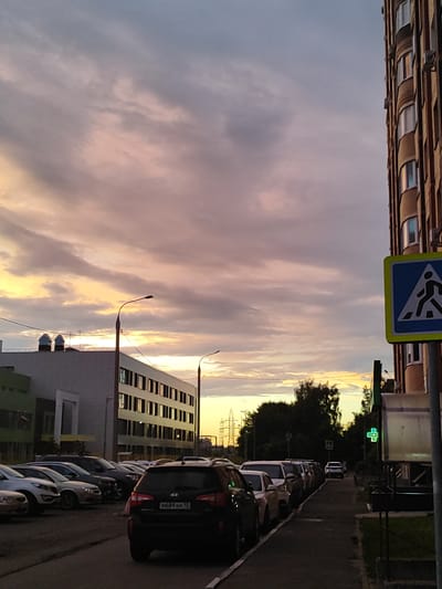 The image shows a street scene at dusk in Shchyolkovo, Russia. The sky is filled with clouds tinged with pink and orange hues from the setting sun. On the right side of the frame, a tall, multi-story apartment building with a brick facade is visible, featuring many windows. A pedestrian crossing sign with a blue background and a white silhouette of a person walking is mounted on a pole in front of the building. To the left of the apartment building, across the street, is a long, multi-story building with a light-colored facade and numerous dark-tinted windows. Several cars are parked along the street. In the foreground, the rear of a black SUV is visible, with its license plate showing "M689AM 15". Further down the street, more cars are parked, and a sidewalk runs alongside them. A green cross symbol, indicative of a pharmacy, is lit up on a pole to the right of the pedestrian crossing sign, behind a structure that appears to be a kiosk or shelter. Trees line the background, and in the distance, some industrial structures and power lines can be seen against the horizon. The overall atmosphere suggests a calm evening in a residential urban area.