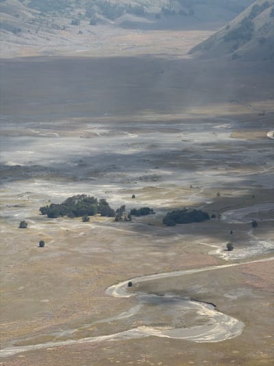 This is a high-angle, long shot of a vast, arid landscape in Cemorolawang, Indonesia. The terrain is characterized by rolling hills and a flat, sandy plain with a winding river or dry riverbed. Sparse vegetation, including small clumps of trees and scattered bushes, dot the landscape. A dirt road snakes through the plain. The scene is bathed in soft, diffused light, with shadows casting long streaks across the ground. The atmosphere appears hazy, possibly due to dust or mist. No people are visible in the image. The environment suggests a remote, undeveloped area with natural geological formations.
