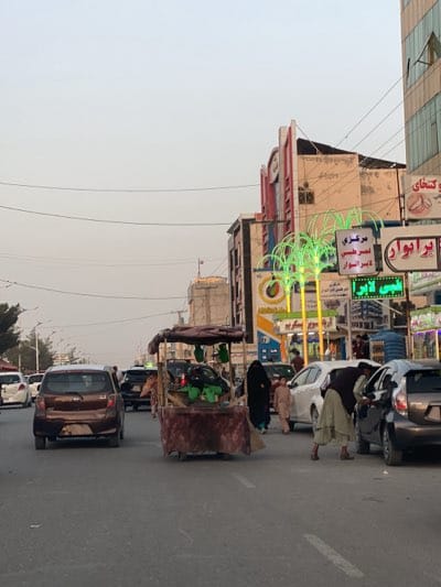 The image depicts a street scene in Kandahar, Afghanistan. The road is populated with several cars, and a vendor's cart is situated in the middle of the road. Several people are present; one person is leaning into a car and another is walking alongside a child. Several businesses are visible on the side of the road with signs written in Arabic. The sky is overcast.