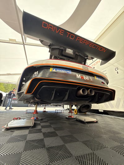 The rear end of a black and white race car is on jack stands in a garage-like setting, on checkered black and grey floor tiles. The rear wing of the car is visible and has the words "DRIVE TO PERFECTION" in red letters. The rear bumper has "MICHELIN" and "Mobil 1" logos. The car is in Stavelot, Belgium (latitude: 50.441, longitude: 5.969). A man wearing a black shirt and jeans is standing by the car, looking at it. He has a cap on. There is a sign behind him that says "ERA CUP HOLLAND". The car's tailpipes are visible. The rear wheels are removed and the car is sitting on jack stands. The underside of the car is also visible. The car is facing towards the right side of the image. The scene appears to be a garage or repair shop. The image is taken from a low angle.