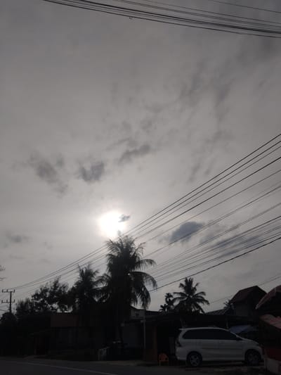 This is a high-angle, medium shot photograph taken in Keudebayu, Indonesia, on a day with overcast skies and diffused sunlight. The scene captures a roadside view with several power lines crisscrossing the frame. In the lower right, a white minivan is parked. To the left of the minivan, several buildings are visible, including one with a reddish-brown roof. Silhouetted against the sky are several palm trees, with the sun appearing as a bright, hazy orb partially obscured by clouds and a palm frond. A utility pole with electrical wires is visible on the far left of the image, and a paved road runs along the bottom. The overall impression is that of a quiet, somewhat rural street scene under a dramatic sky.