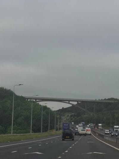The image captures a view from a highway in Kirklees, United Kingdom, under a gray, overcast sky. A multi-lane highway stretches forward, with traffic including cars, vans, and trucks moving in the same direction. On the left side of the highway, a dense line of green trees and foliage runs alongside a steep embankment. Towering over the trees and highway is a large concrete bridge, characterized by its arched structure. Streetlights are visible lining the side of the highway, spaced at intervals, their lamps obscured by the dim lighting conditions. The overall atmosphere is muted due to the cloudy weather, contributing to a somewhat somber or tranquil mood.