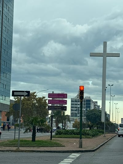 The image depicts a street scene in Montevideo, Uruguay. A large, white cross is prominently visible in the background. Street signs indicate directions to "Hospital Italiano," "Hospital Británico/Clínicas," "Estadio Centenario," "Tnal de Omnibus Tres Cruces," "Plaza de la Democracia," and "Monumento a la Bandera." The traffic light is red. The sky is overcast. There are buildings and trees in the background. On the sidewalk, one individual is walking and another one is standing behind. A palm tree is planted along the sidewalk. The image is taken from a vehicle moving along a road.