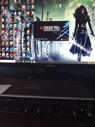 The image shows a computer monitor displaying a desktop with various icons and a Cubase Pro 9 software interface. The desktop background is an image of a woman in a dark, flowing outfit, possibly a fantasy character. The Cubase Pro 9 interface is a black box with white text, showing the version number and other details. To the right of the monitor, a Behringer audio interface is visible, with several knobs and buttons. The room appears to be dimly lit, suggesting it might be evening or indoors. The overall scene suggests a creative workspace, likely for music production, given the software and audio interface. Visible text includes "CUBASE PRO 9 Advanced Music Production System," "Version 9.0.0 (64 bit) - Built on Nov 3 2016," "Initializing: Content," and "Behringer." There are also many application icons on the desktop, indicating a user who has installed and uses a variety of software. The monitor itself is identified by the "PHILIPS" logo. The image is oriented sideways.