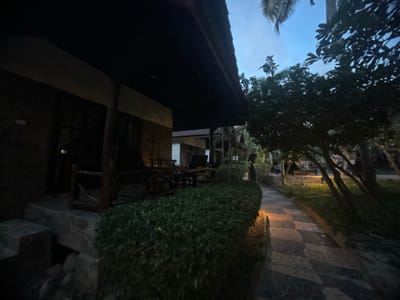 The image shows a stone-paved path in Baan Wang Ta Kien, Thailand, lined with trees and a manicured hedge. To the left, a dark building with a porch is visible. The time of day appears to be dusk or early evening based on the low light levels. No people are visible in the frame. The environment seems tranquil and well-maintained.