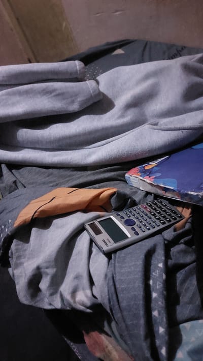 The image shows a close-up, dimly lit, high-angle view of a bed with various clothing items and a calculator. A grey hoodie is draped across the top of the frame, with its sleeves visible. Beneath the hoodie are layers of grey and blue fabric, likely sheets or blankets, with a patterned design on some of the fabric. In the lower right quadrant, a calculator with a silver casing and a green display is resting on the fabric. To the right of the calculator, a book with a blue cover adorned with colorful floral patterns is partially visible. A folded piece of orange fabric with black lines is positioned to the left of the calculator. The background appears to be a textured wall or surface. The scene suggests a domestic setting, likely a bedroom. The image does not provide any specific details to indicate a location in the city of New Nyanya, Nigeria.