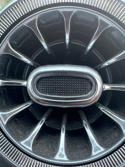 This is a close-up, eye-level shot of a chrome-plated car air vent. The vent is circular with radiating fins and a central, oval-shaped intake with a textured black insert. The chrome is slightly dulled by a layer of dust, which is also visible on the black textured surface and in the recesses of the fins. The background is dark, suggesting the interior of a vehicle. There is no indication of the city of Taldom, Russia, or any people.