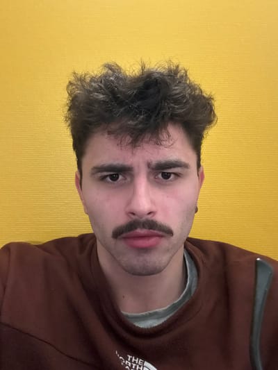 A young man with dark hair and a drawn-on mustache poses for a selfie. He wears a brown sweatshirt over a gray undershirt. Part of a pair of glasses is visible at the top edge. The background is a plain yellow wall. The image is oriented sideways.