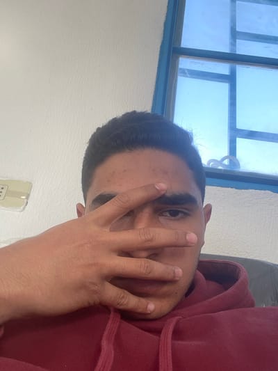 A man in a maroon hoodie partially covers his face with his hand, obscuring most of his features. One eye is visible, gazing towards the camera. He has short, dark hair. Behind him, a window with blue frames allows natural light into the room. The wall is a plain white. A small, beige electrical outlet is partially visible in the lower left corner of the image. The picture is taken at an angle, with the subject appearing sideways.
